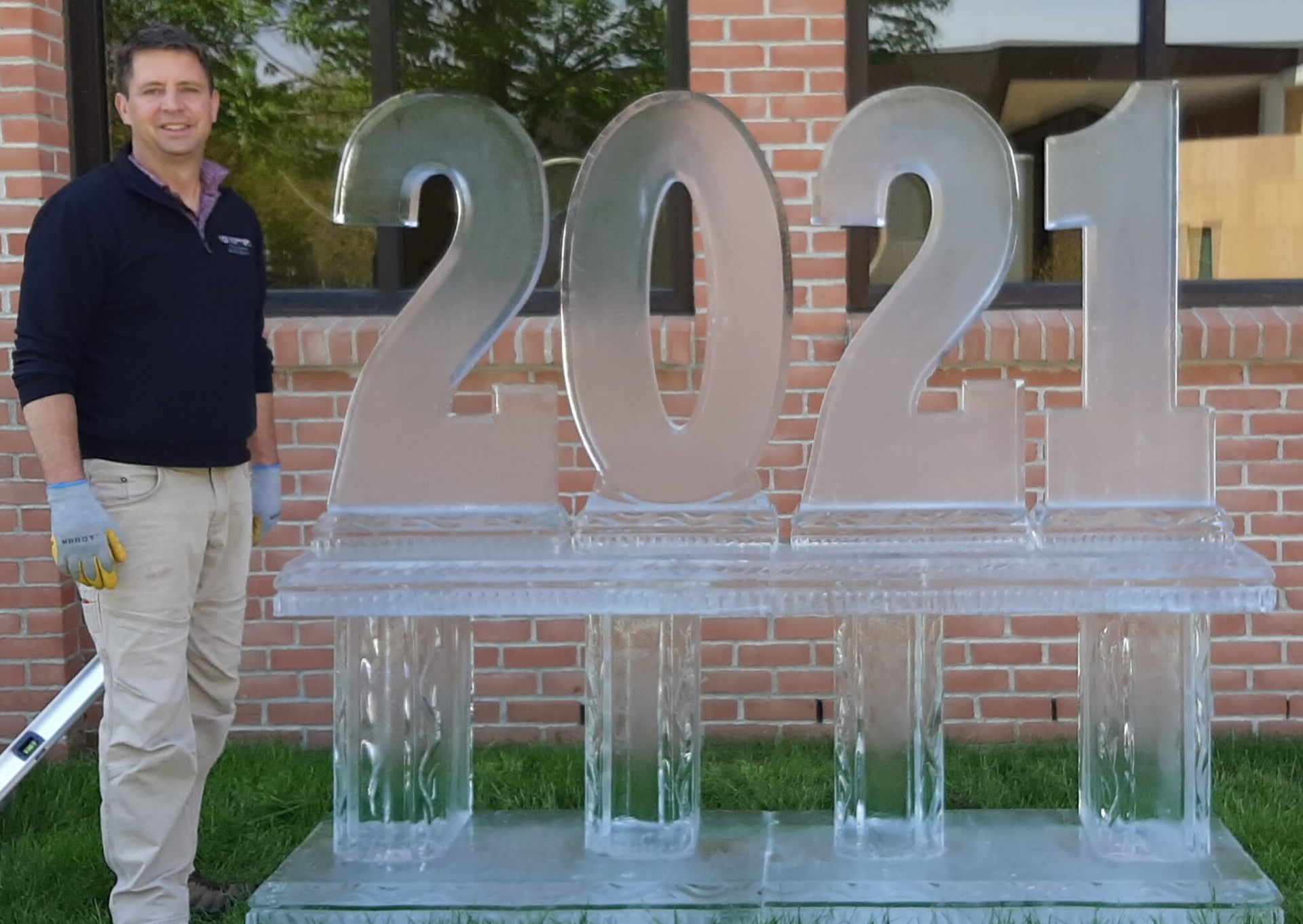 Man standing beside a large clear ice sculpture reading “2021” in front of a brick building.
