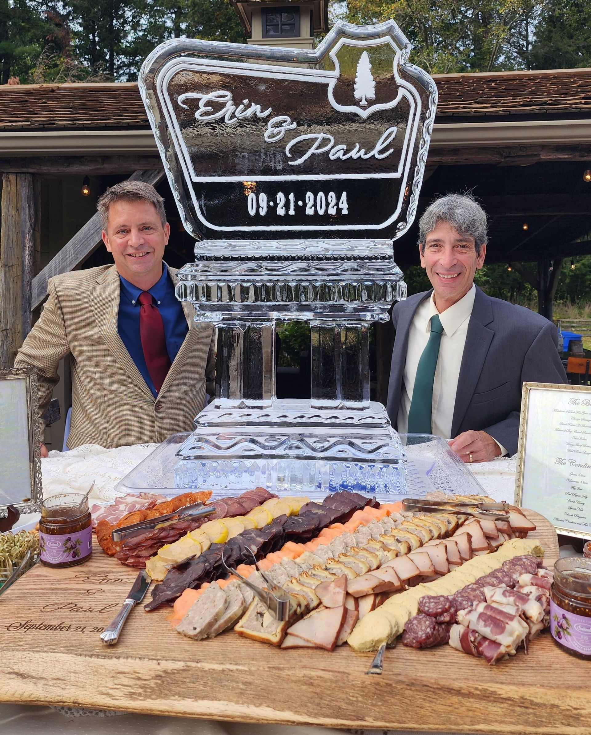 Two men posing behind a large meat platter and an ice sculpture with a date plaque outdoors