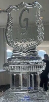 Clear glass trophy with a large letter “G” on top and a tiered base, displayed indoors.