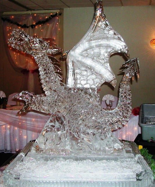 A clear ice sculpture of a dragon displayed indoors on a pedestal, with a white sail-like shape behind it.