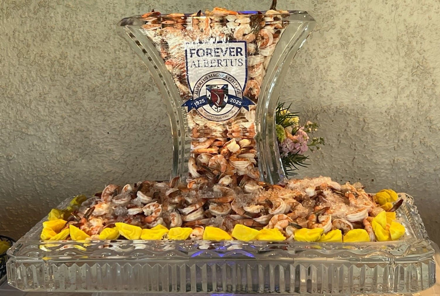 Glass tray of seashells with yellow flowers, centered around a decorative labeled vase on a beige wall background