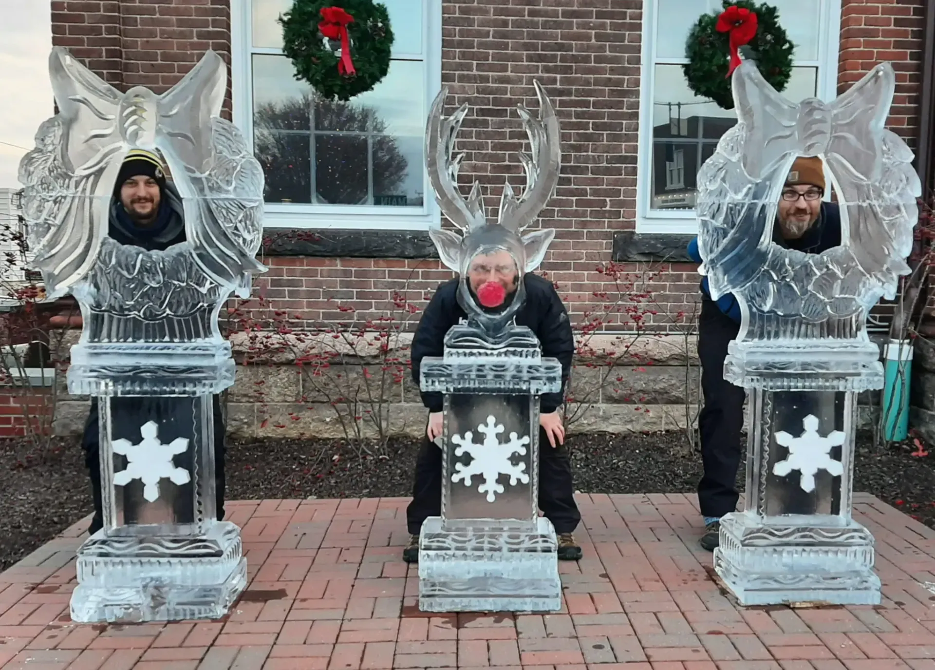 Three people posing in icy themed cutout displays on a brick sidewalk with holiday wreaths behind them.