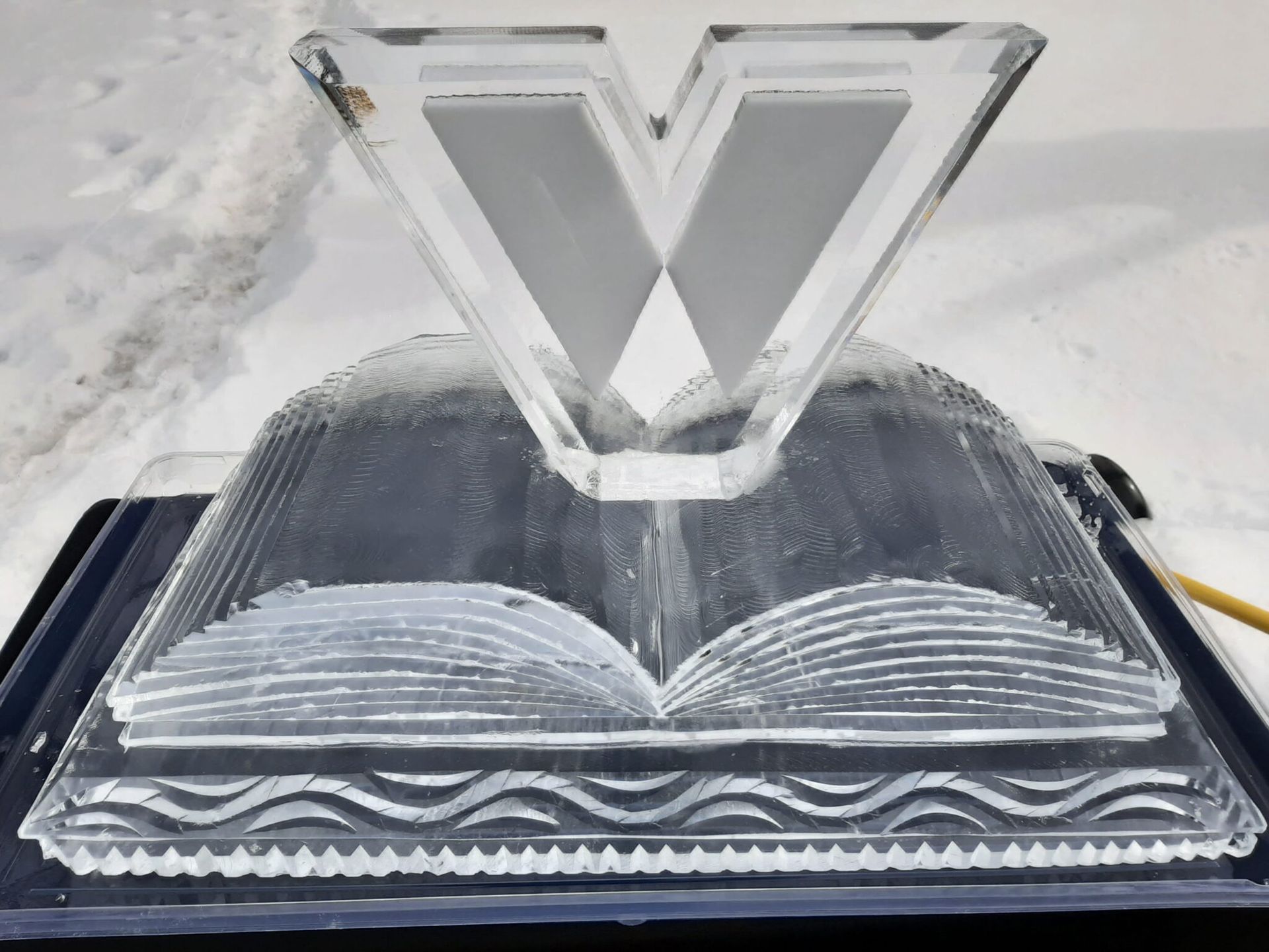 Transparent V-shaped sculpture standing on an open book, on a snowy white surface