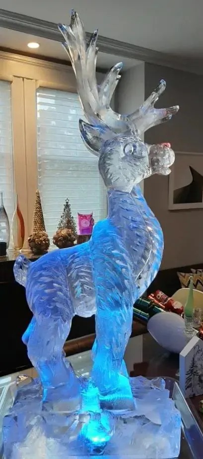 Blue-lit clear glass reindeer sculpture with antlers on a table indoors, beside holiday decor.