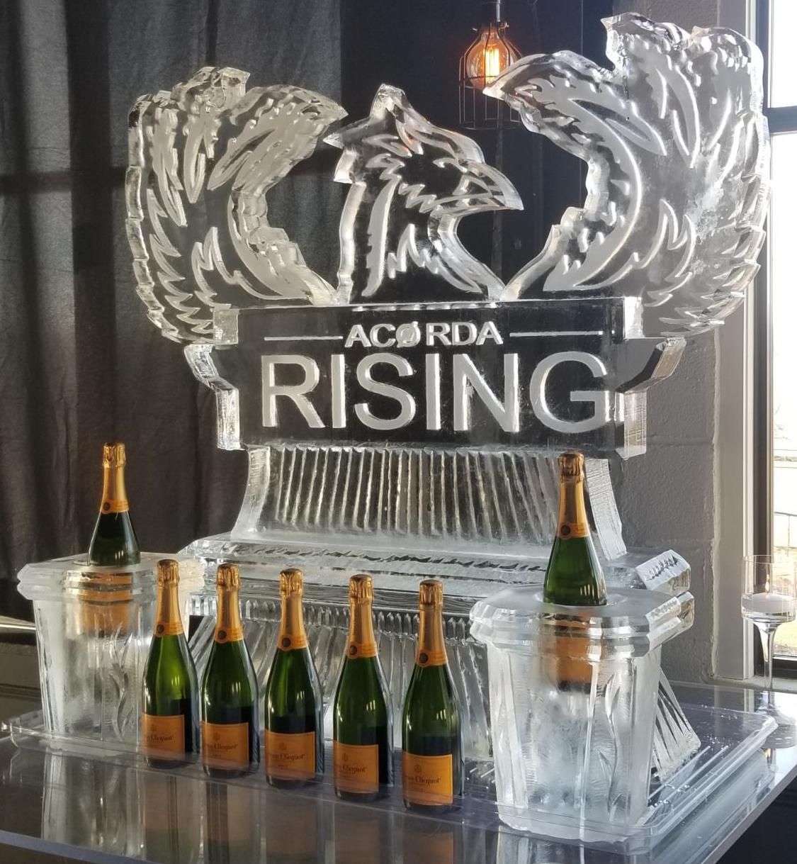 Ice sculpture reading “AC RDA RISING” with champagne bottles on a table in front of gray curtains