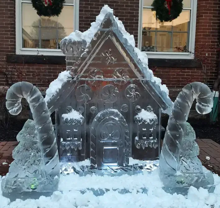 Ice sculpture of a gingerbread house with candy cane swirls, on snow outside a brick building.