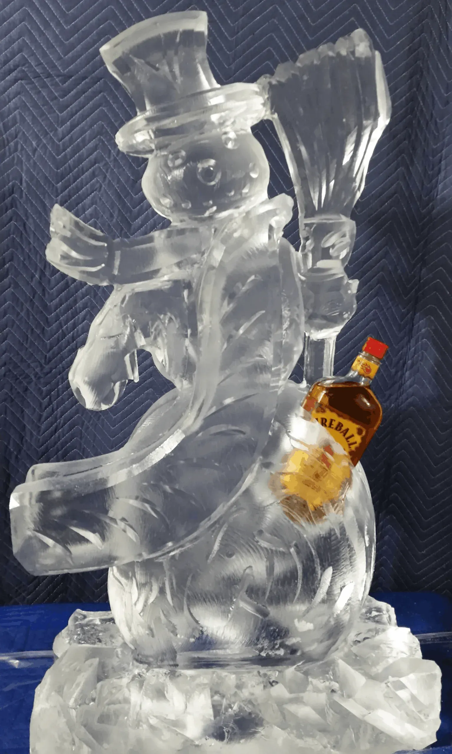 Clear ice sculpture of a magician holding a wand, with a small amber bottle beside it on a dark blue background