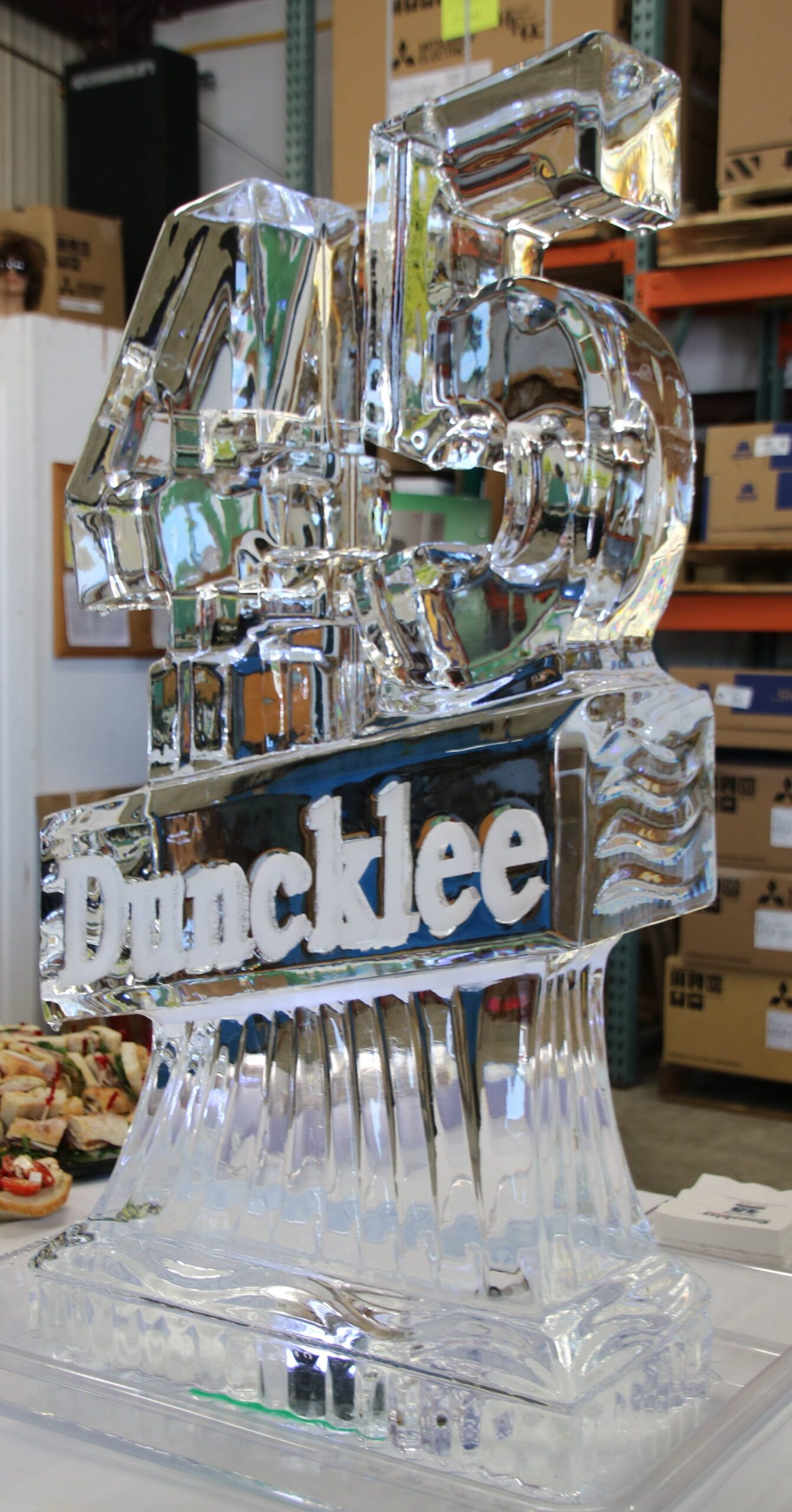 Clear ice sculpture of a large “M” logo on a base at an indoor event display.