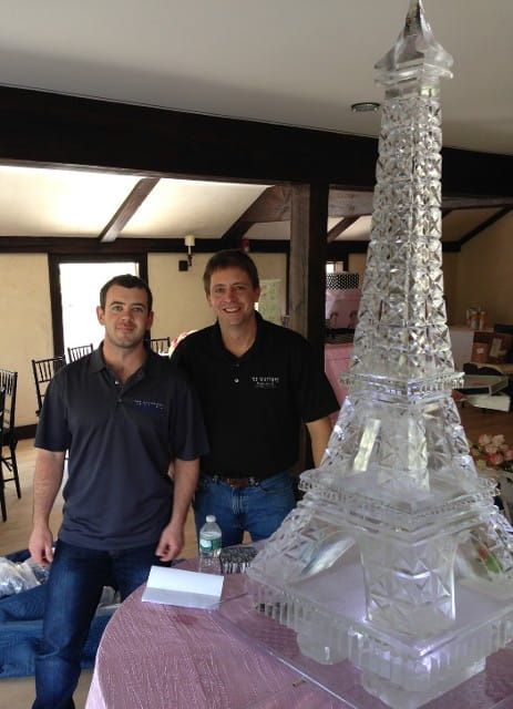 Two men standing beside a tall clear glass sculpture in a room with exposed wooden beams