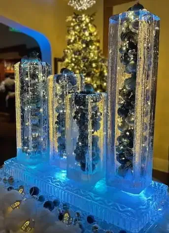 Blue-lit glass towers with silver ornaments, arranged on a decorated holiday table beside a lit Christmas tree.