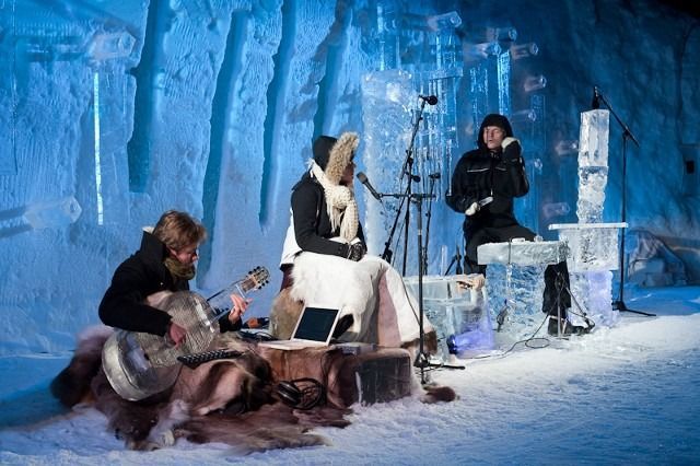 Three people in winter clothing sit in an icy blue cave-like setting with sculptures and a small fire.