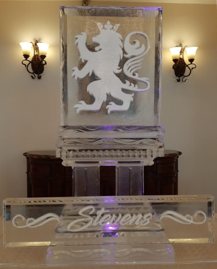 Clear ice sculpture with a lion crest and the name “Stevens” etched on a block, indoors with wall sconces.