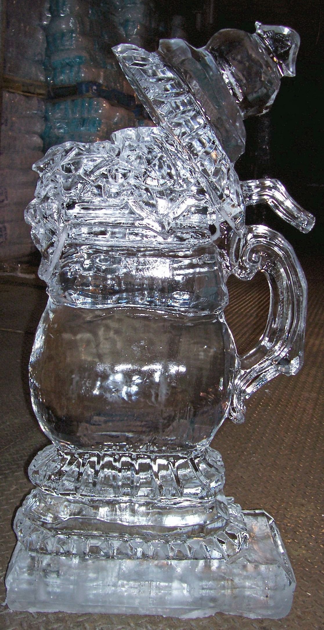 Ice sculpture of a glass pitcher with handle and lid on a base, shown indoors against a dark background