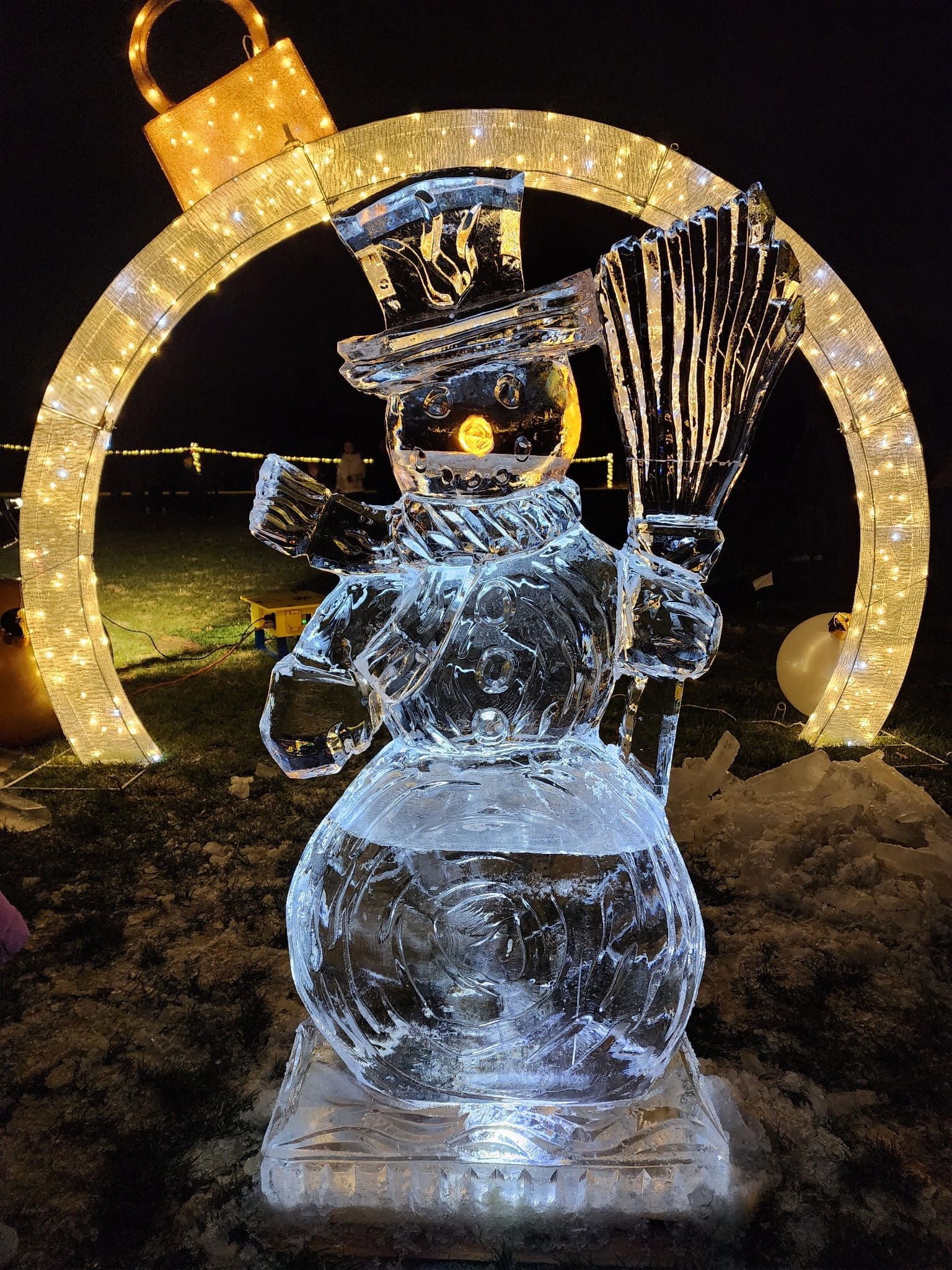 Snowman ice sculpture with a top hat and broom, lit up at night in front of a glowing arch.