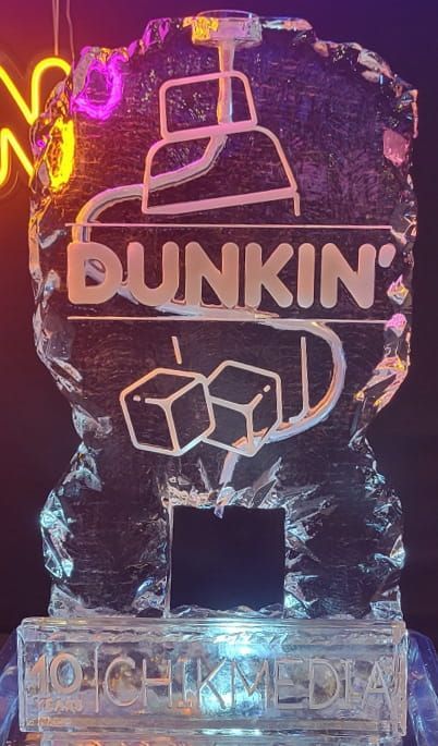 Ice sculpture of a Dunkin' coffee cup logo, lit in pink and purple neon, with ice cubes on a pedestal.