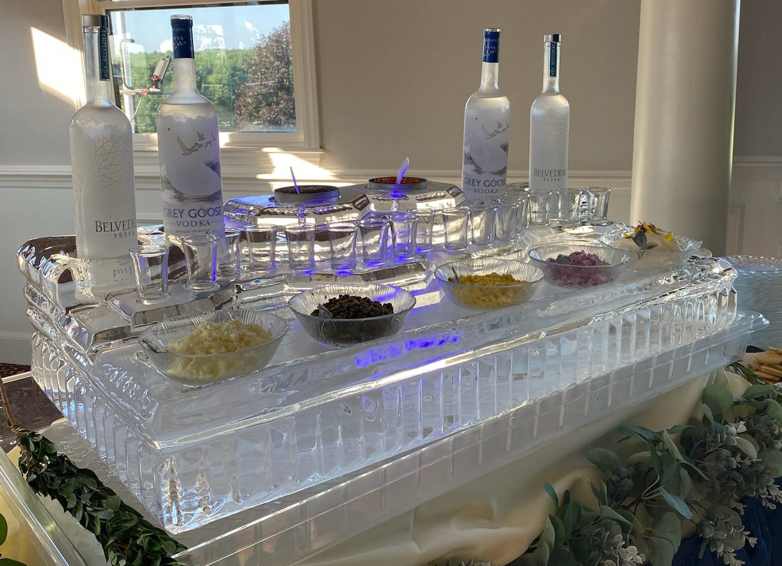 Ice bar display with liquor bottles, glasses, and small bowls on a reflective tray outdoors