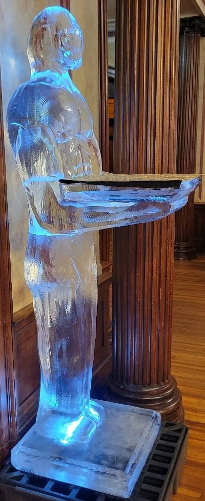 Blue-lit transparent ice sculpture of a person holding a tray, indoors beside a wooden column.