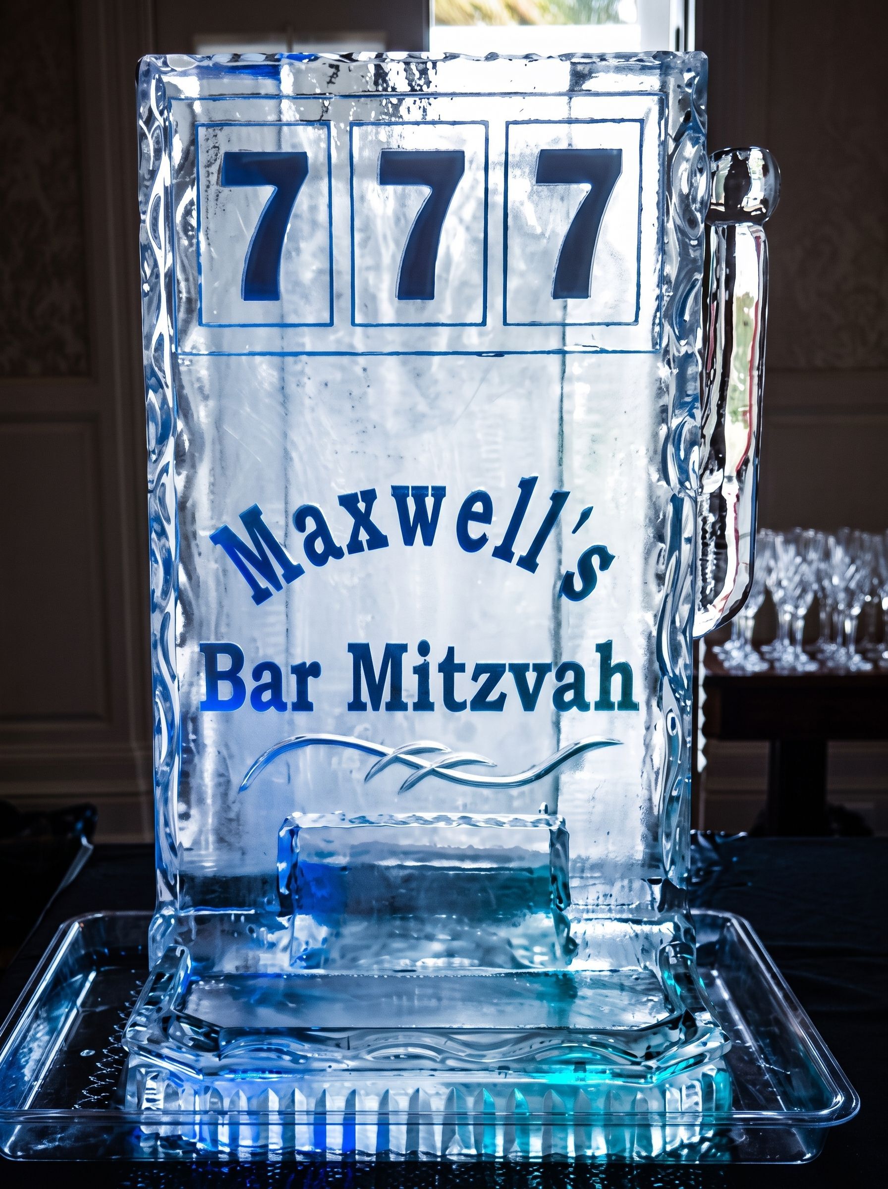 Blue ice sculpture reading “Maxwell’s Bar Mitzvah” with three 7s on top on a table.