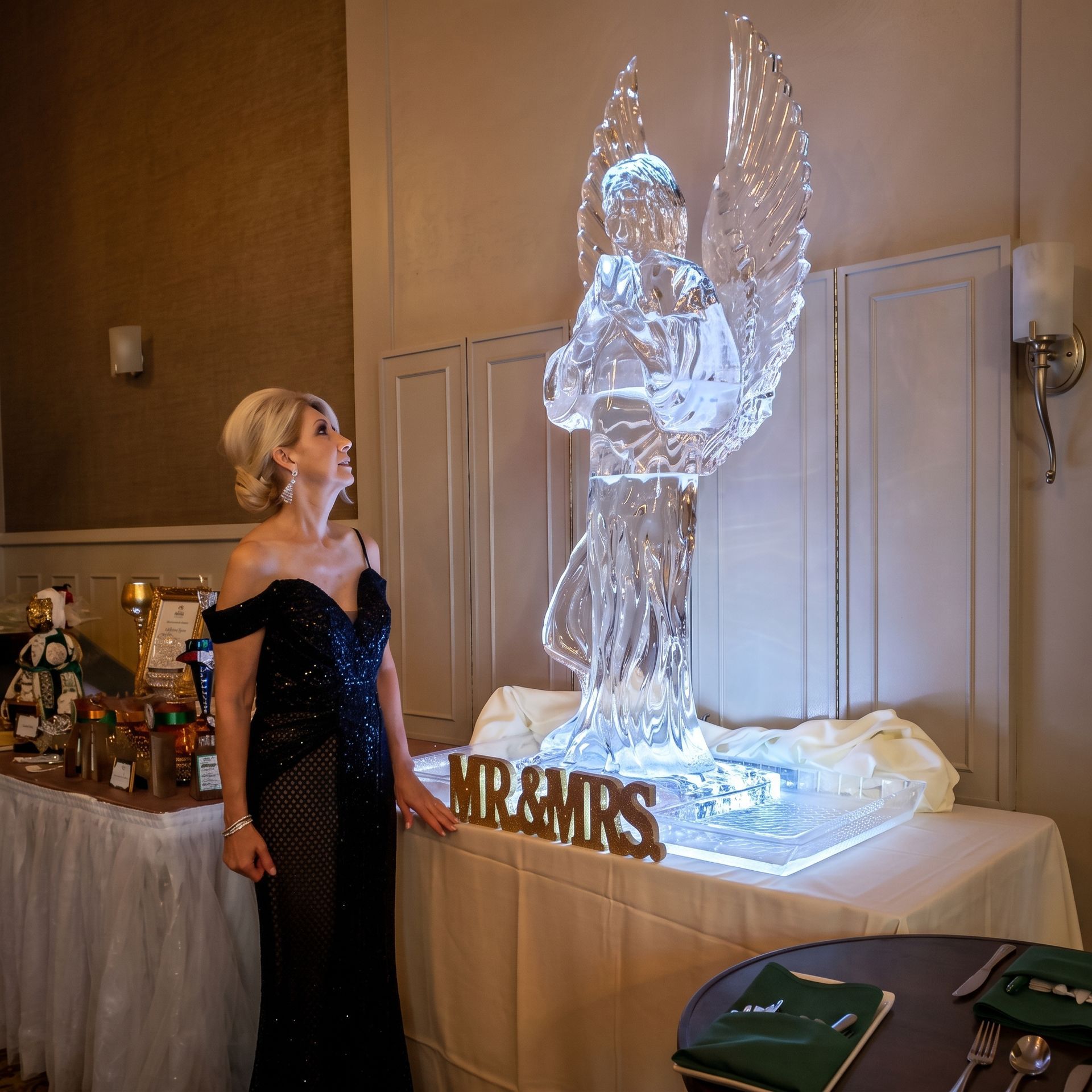 Woman in a black gown admiring a glowing ice angel sculpture at an indoor event table