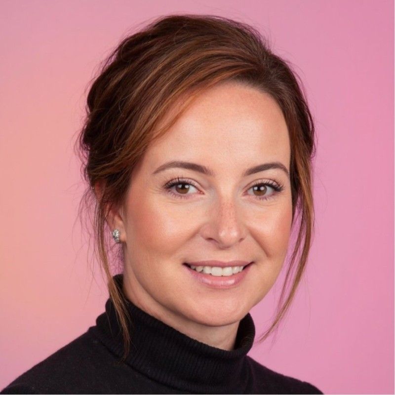 Woman with brown hair smiles, wearing a black turtleneck, set against a pink gradient backdrop.