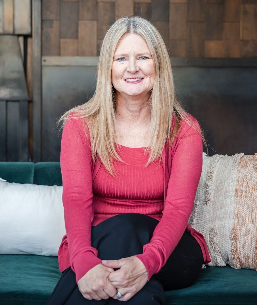 Woman with blonde hair wearing a red top and black pants, sitting on a teal couch.