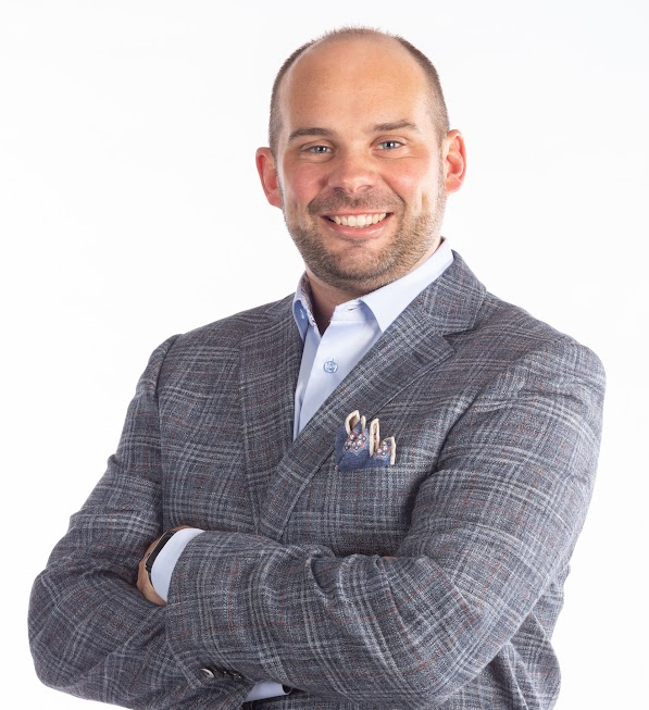 Man in a plaid blazer and open-collared blue shirt, arms crossed, smiling.
