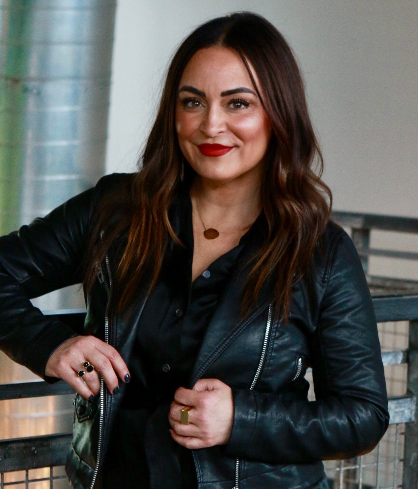 Woman in black leather jacket and dress, red lipstick, leaning on a railing.