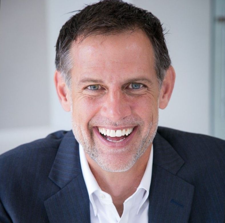 Man in a navy blazer and white shirt smiles widely, blue eyes, neutral background.