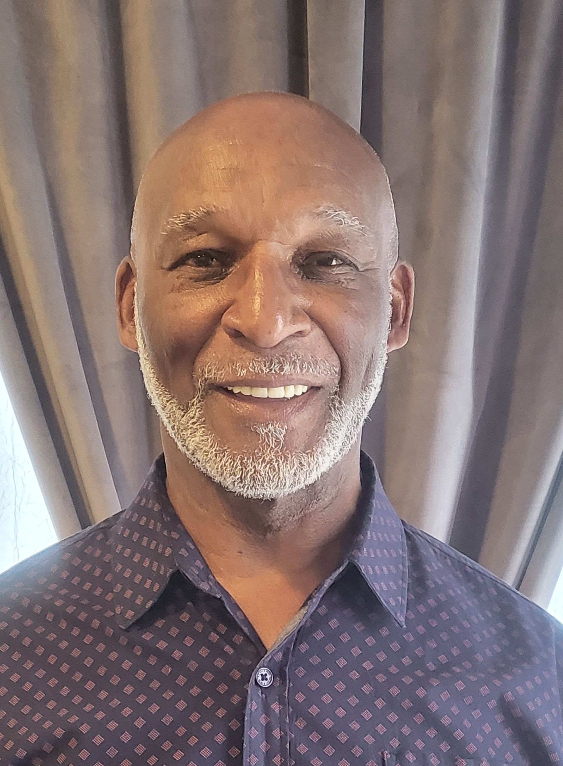 Smiling man in a navy patterned shirt standing before gray curtains