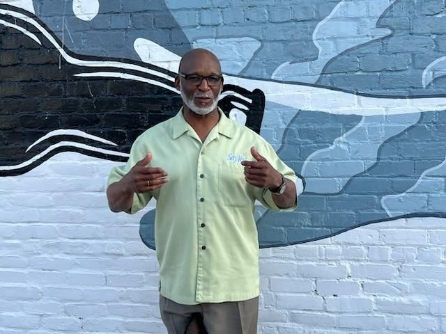 Man in light green shirt gesturing in front of a blue-gray mural wall