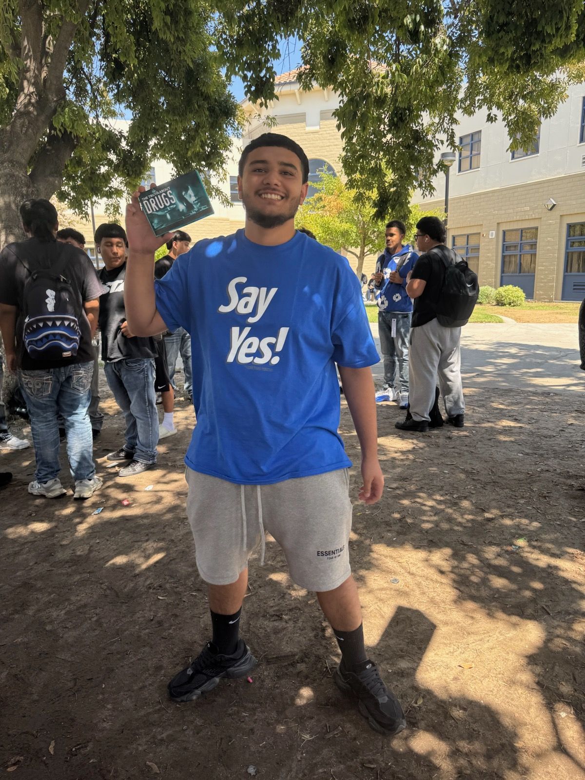 Person in a blue “Say Yes!” T-shirt stands outdoors, smiling and holding a cup near a building.