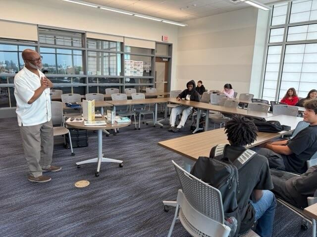 Presenter speaking to seated group in a modern classroom with tables and large windows