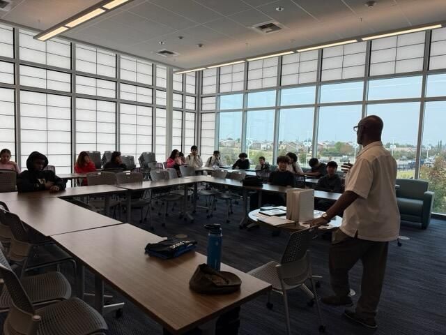 People seated around conference tables in a bright meeting room with large windows; one person stands presenting.