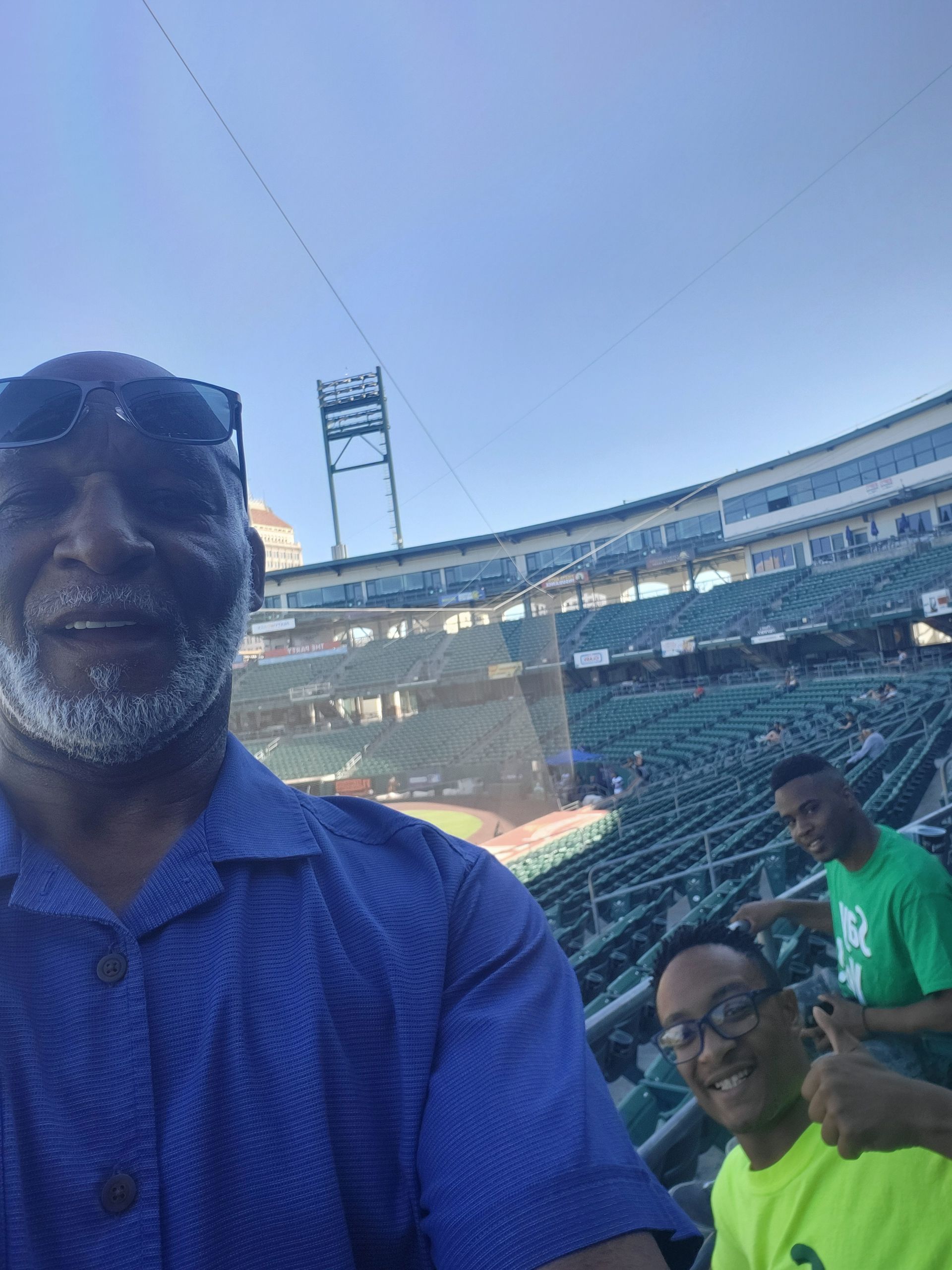 Man taking a selfie in a stadium with green seats and two people in the background.