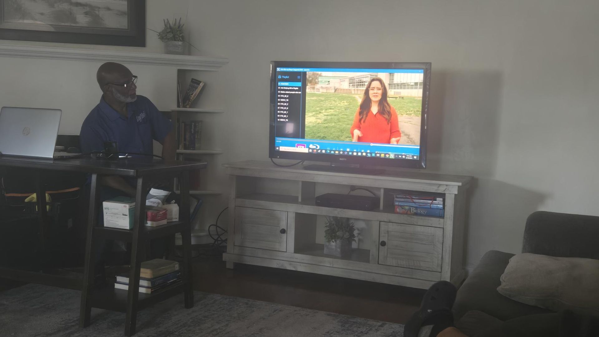 Living room with a person seated beside a TV showing a news broadcast