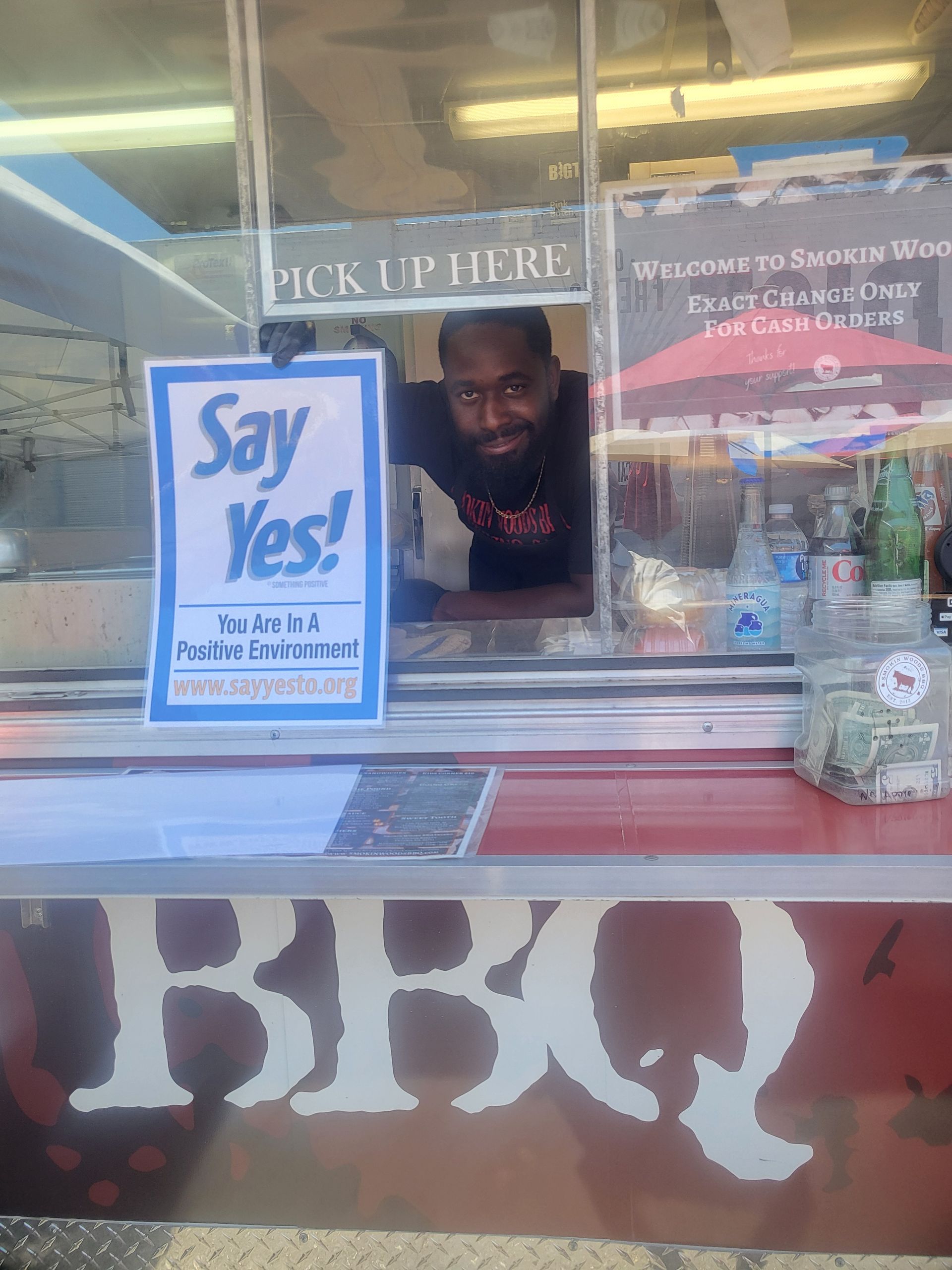 Man leaning out of a BBQ takeout window beside a blue “Say Yes!” sign.