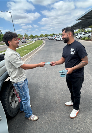 Two men exchange an item on a street beside parked cars, under a cloudy sky.