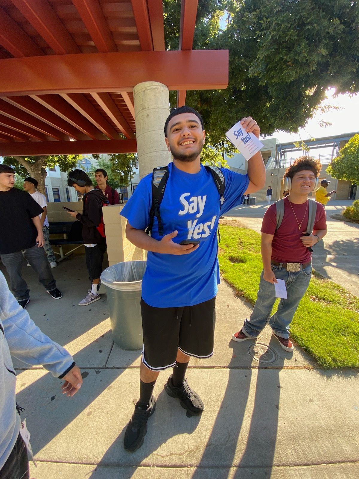 Smiling person in a blue “Say Yes” shirt holding a flyer at an outdoor event under a pavilion