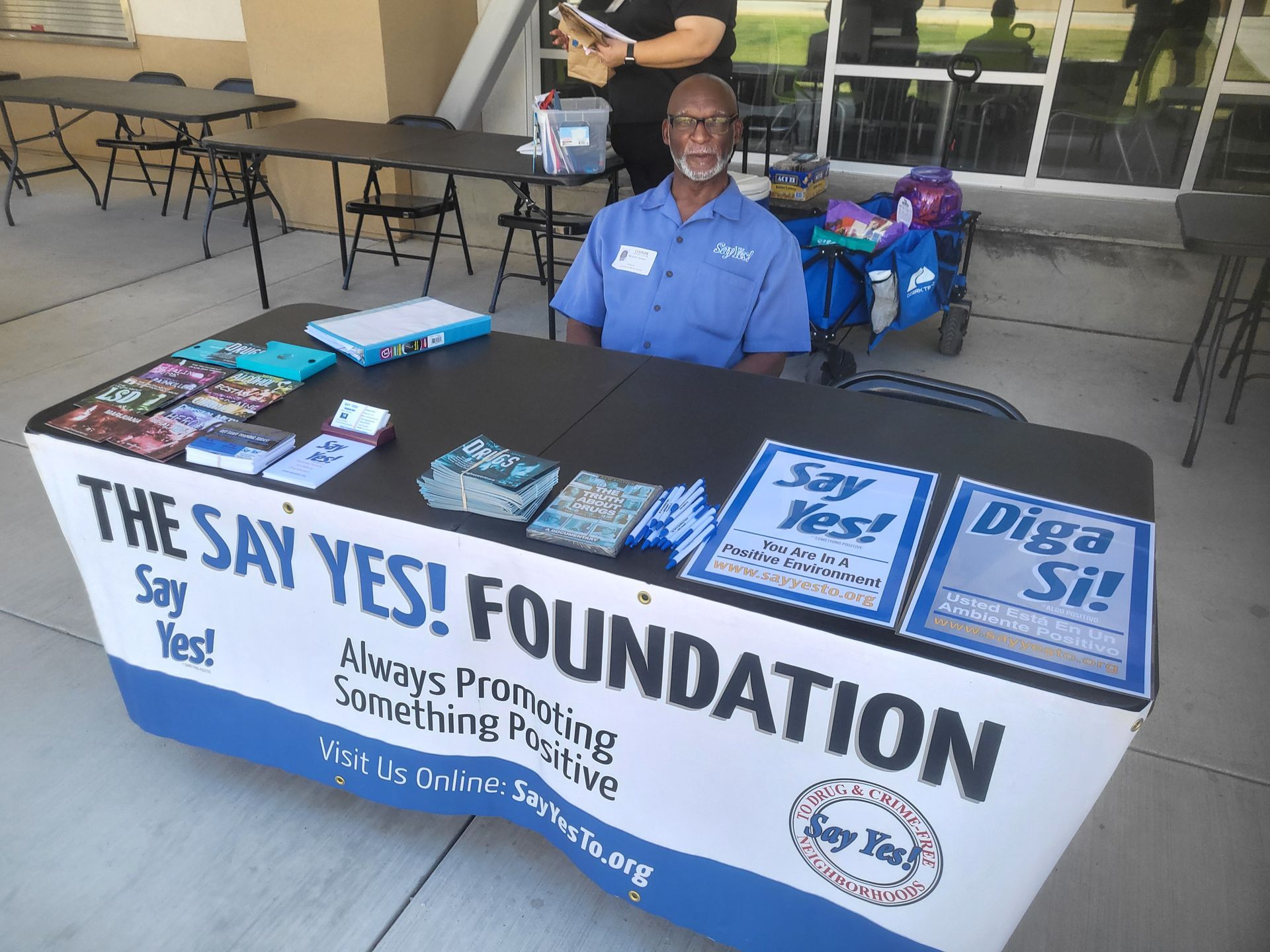 Volunteers at a SAY YES! Foundation table with brochures and a smiling representative seated under a canopy.
