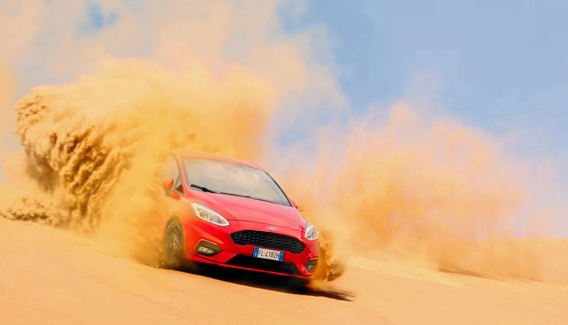 Red car speeding through sand, kicking up a large cloud, under blue sky.