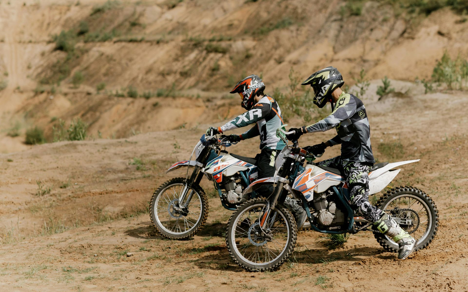Two people on dirt bikes on a sandy track, wearing helmets and protective gear.