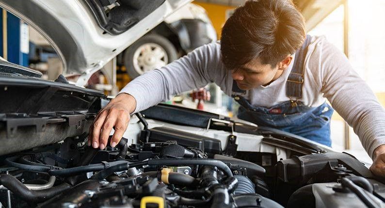 Mechanic working under an open car hood in a garage, inspecting engine components.
