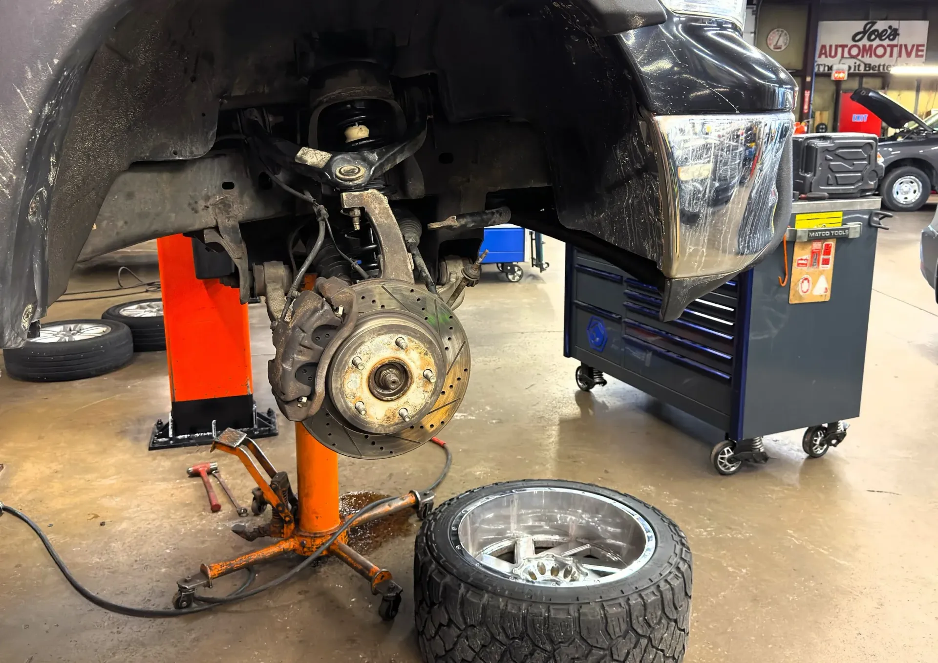 Car in auto shop with rear wheel removed, exposing brake assembly; tire lies on the floor nearby.