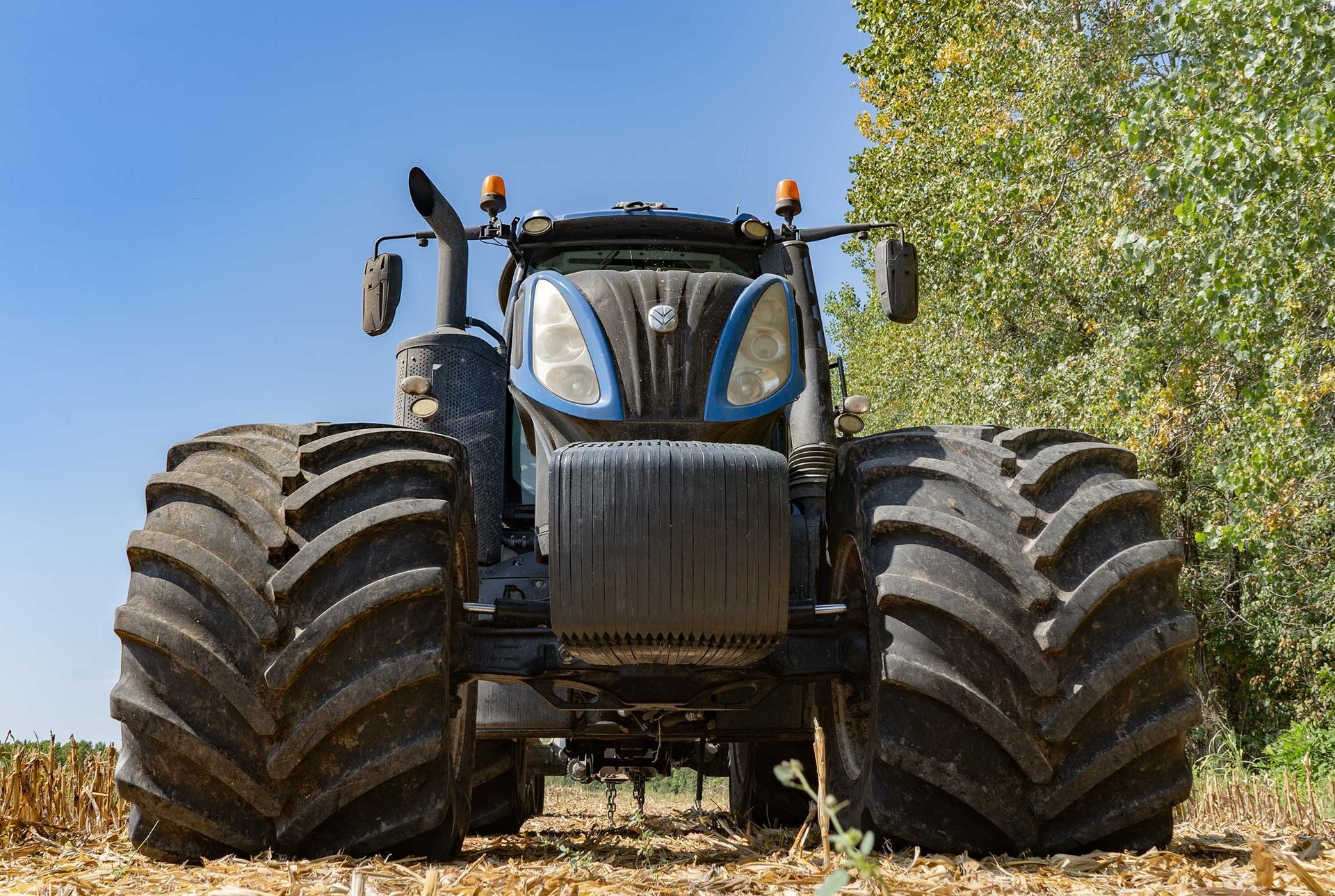 Front view of a large black tractor with oversized tires on a dirt field beside a tree