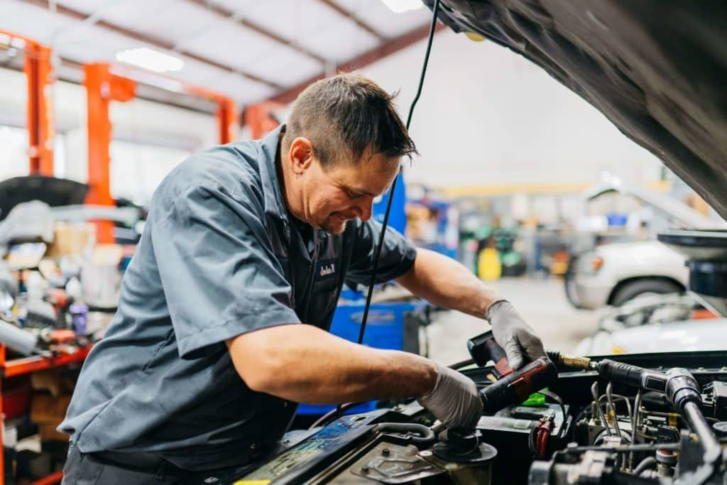 Mechanic in gray shirt working under a car hood in an auto repair shop