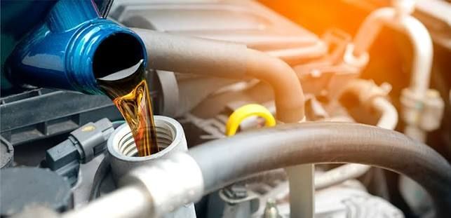 Blue oil being poured into a car engine through the filler cap, with hoses and engine parts visible.