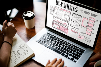 Mani che abbozzano wireframe di un sito web accanto a un laptop che mostra layout dell’interfaccia utente e appunti