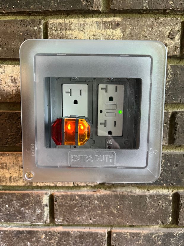 Outdoor wall outlet with two sockets in a clear cover; left socket has an amber plug-in light, right socket has a green indicator.
