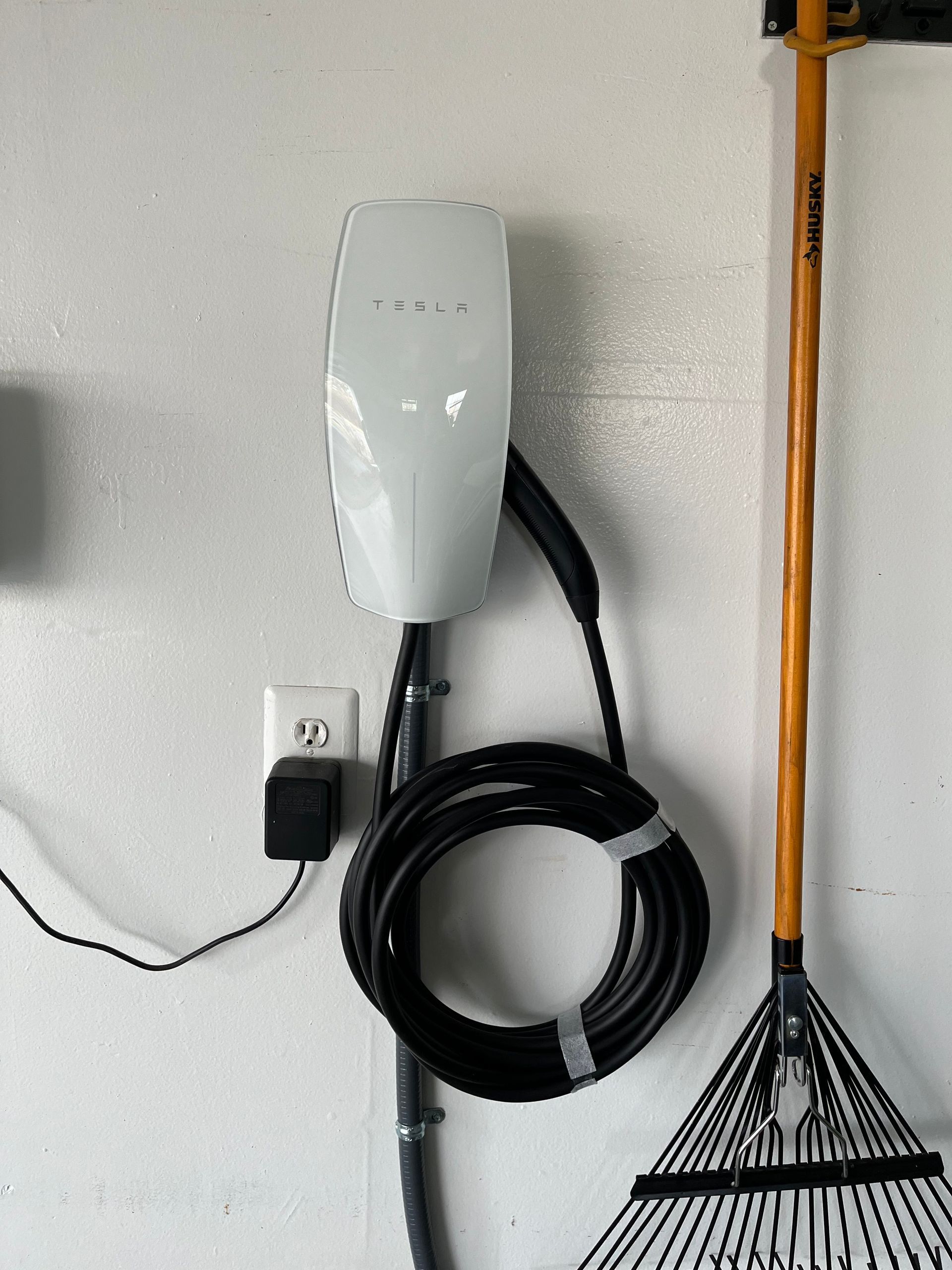 Wall-mounted electric vehicle charger with coiled cable beside a rake on a white garage wall