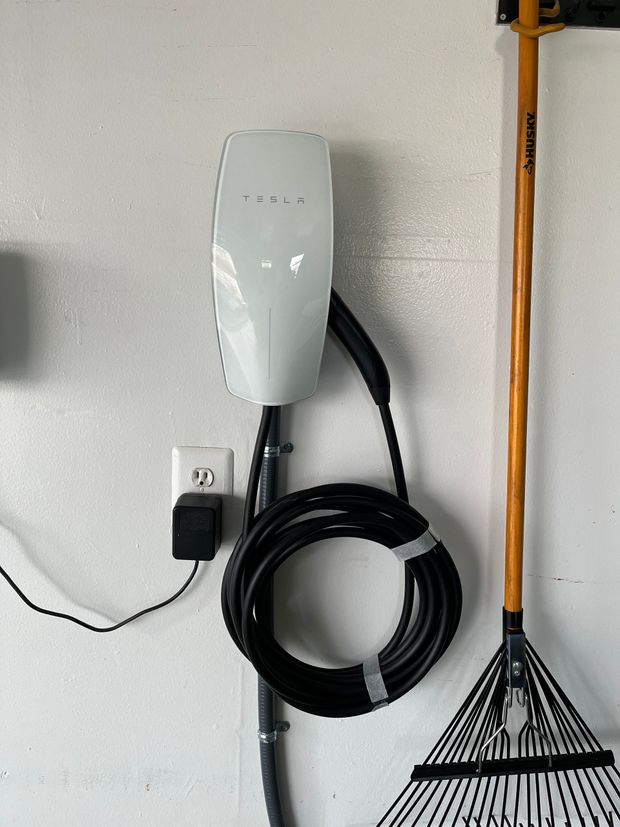 Wall-mounted electric vehicle charger with coiled cable beside a rake on a white garage wall